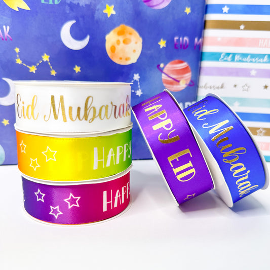 Stack of colorful satin ribbons with 'Eid Mubarak' and 'Happy Eid' text in gold and silver foil, against a background of gift wrap with moon and stars design.
