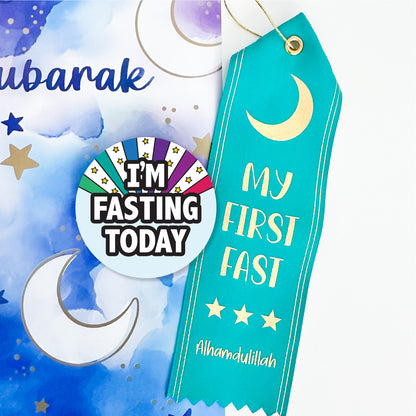 A collection of Ramadan-themed items including a round pin-back button with the text 'I'M FASTING TODAY' and a turquoise satin ribbon award with the text 'MY FIRST FAST' and a crescent moon design.