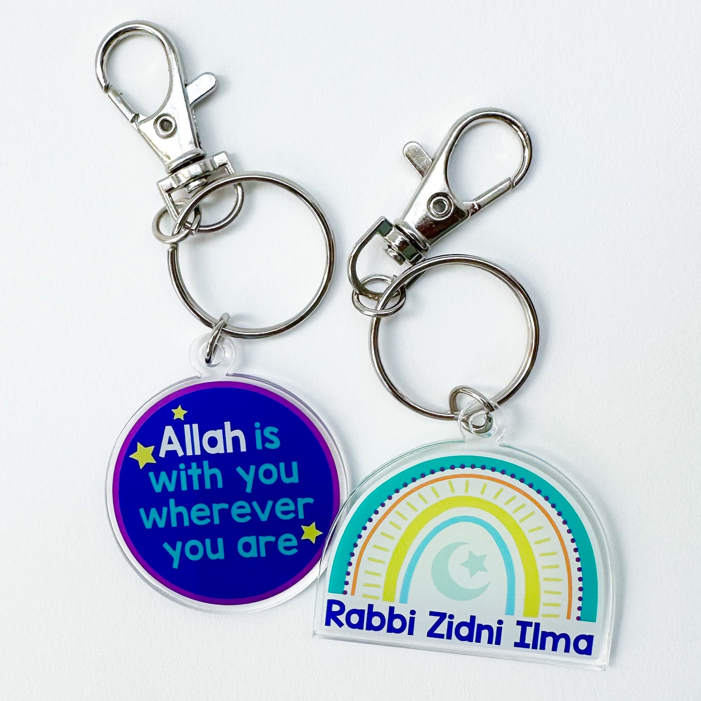 Two acrylic keychains with one being circular with text and the other being rainbow-shaped with a similar text. Both have metal clasps and keyring attachments.